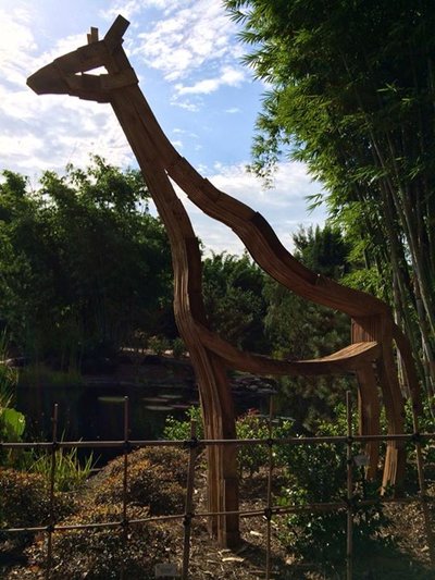 The lifesize stylized rough plywood giraffe overlooks a pond at the zoo.
